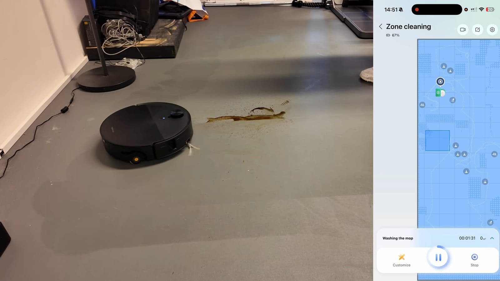 Zwarte robotstofzuiger rijdt naar een plas koffie op een grijze vloer, rechts de app met zoneschoonmaak